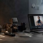 Birou de lucru creativ cu aparat foto analog, cameră profesională și laptop care afișează un portret editat, simbolizând trecerea de la fotografia clasică la producția vizuală AI.
